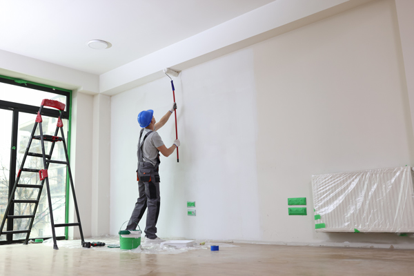 person painting a wall in a room using a paint roller while standing on a ladder with painting supplies nearby and a fresh coat of paint