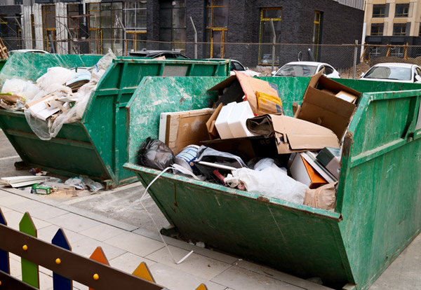 Two green dumpsters filled with assorted waste and construction debris in an urban setting with visible cars and fencing nearby showing the importance of recycling and waste management