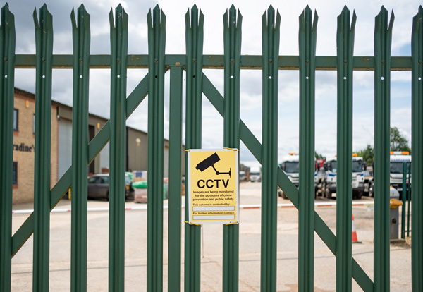 yellow CCTV warning sign on green metal gate at commercial property with parked vehicles in the background providing surveillance security information