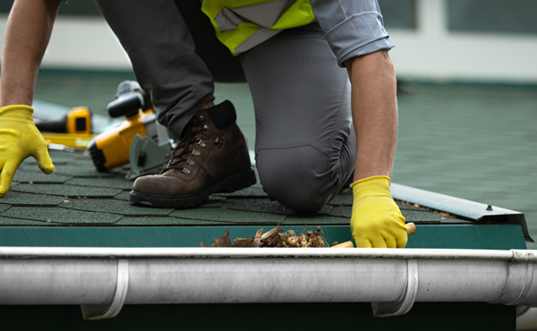 Worker in gloves cleaning debris from roof gutter with tools nearby maintaining home safety and roof health